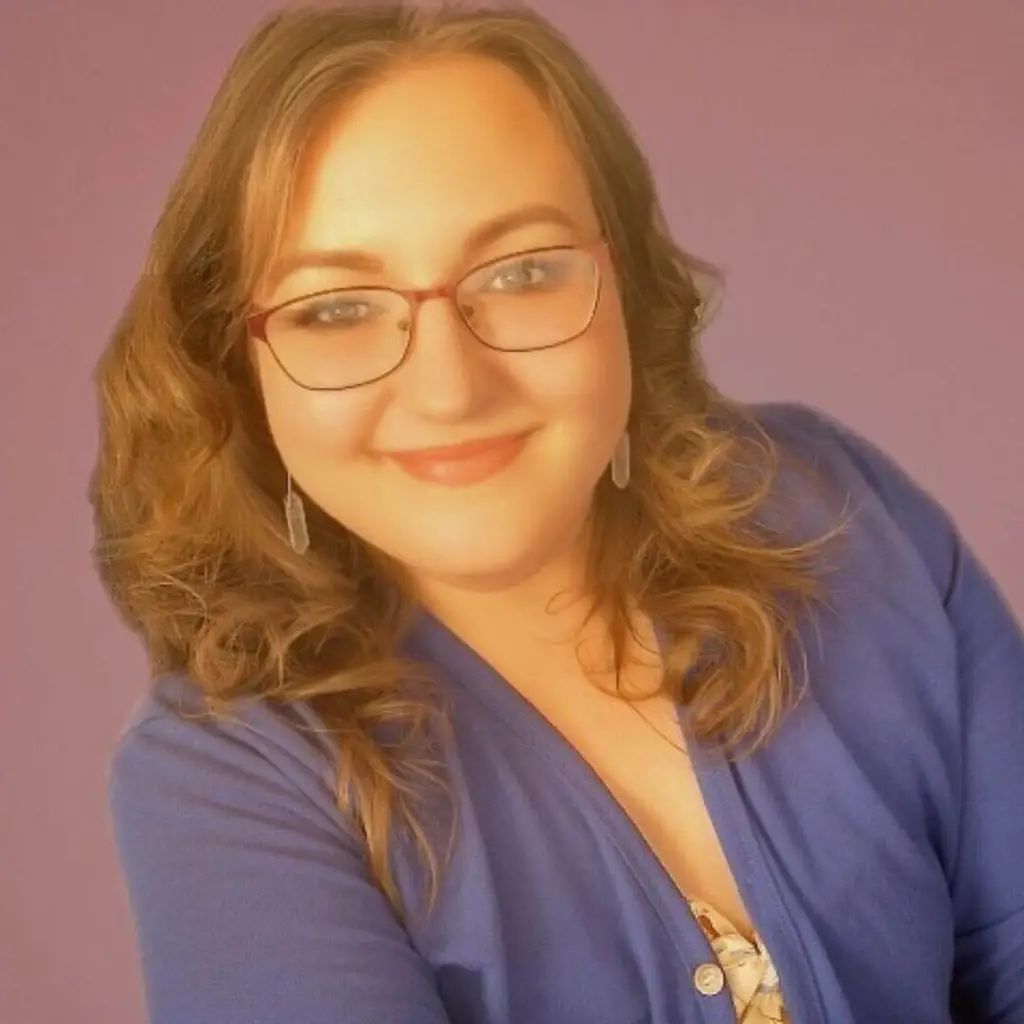 Una mujer con el pelo castaño claro ondulado, gafas y una sonrisa confiada posa delante de un fondo morado, con un top azul.