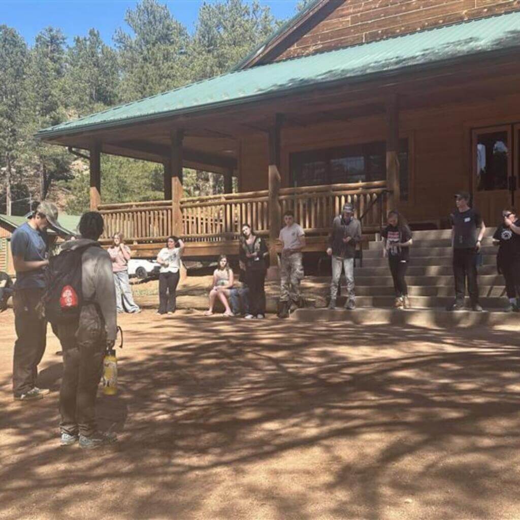 Un grupo de jóvenes se sienta junto a troncos y bancos en una zona boscosa a la sombra, charlando y riendo despreocupadamente cerca de una cabaña de madera.