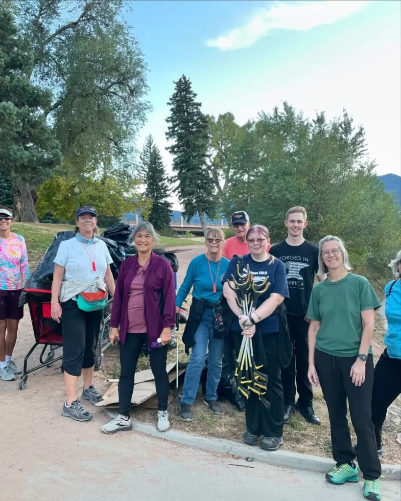 Un grupo de diez personas sonriendo al aire libre, sosteniendo herramientas de jardinería, cerca de bolsas de basura recogidas. Están rodeados de árboles altos y un cielo despejado, lo que transmite un mensaje de trabajo en equipo y servicio comunitario.