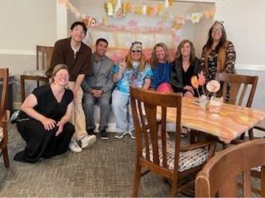 Un grupo de siete personas, sonrientes y sentadas, en una sala decorada con guirnaldas florales y en tonos pastel. El ambiente es festivo y alegre.