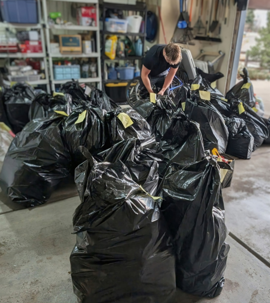 Una persona se inclina sobre un gran grupo de bolsas de basura negras llenas en un garaje, colocando o revisando las etiquetas de las bolsas. La pared trasera está llena de estanterías con artículos de almacenamiento.