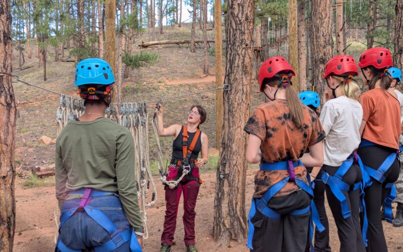 Un grupo de personas con cascos y arneses se encuentra entre altos pinos mientras escucha a un instructor que les muestra cuerdas y equipo de escalada en un curso de aventura al aire libre.