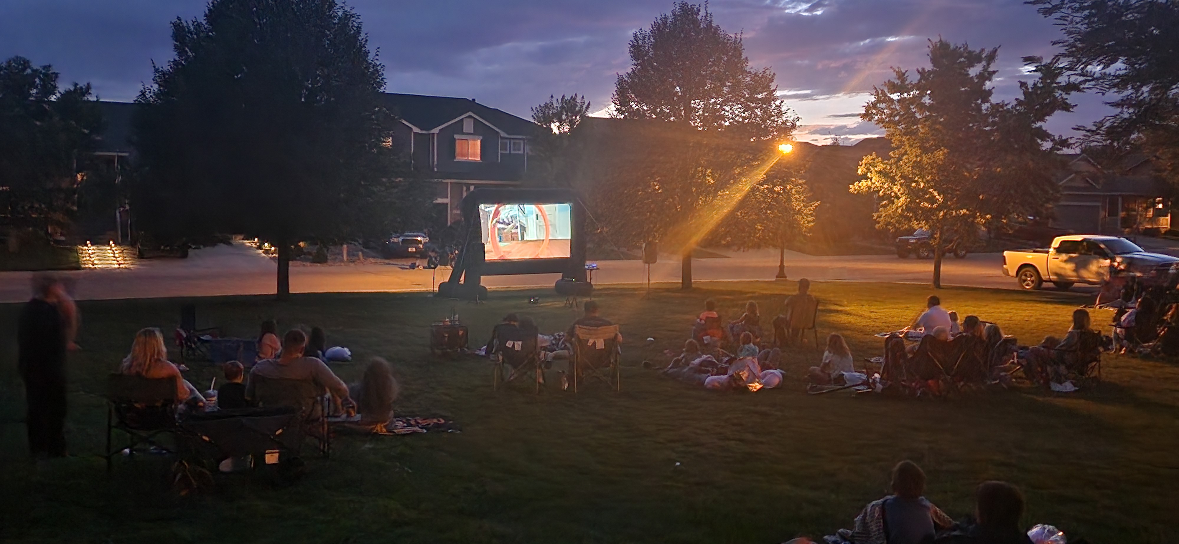 Las familias y los miembros de la comunidad se sientan en mantas y sillas de jardín en un parque cubierto de césped al atardecer, viendo una película proyectada en una gran pantalla al aire libre, mientras las farolas y las casas cercanas brillan al fondo.